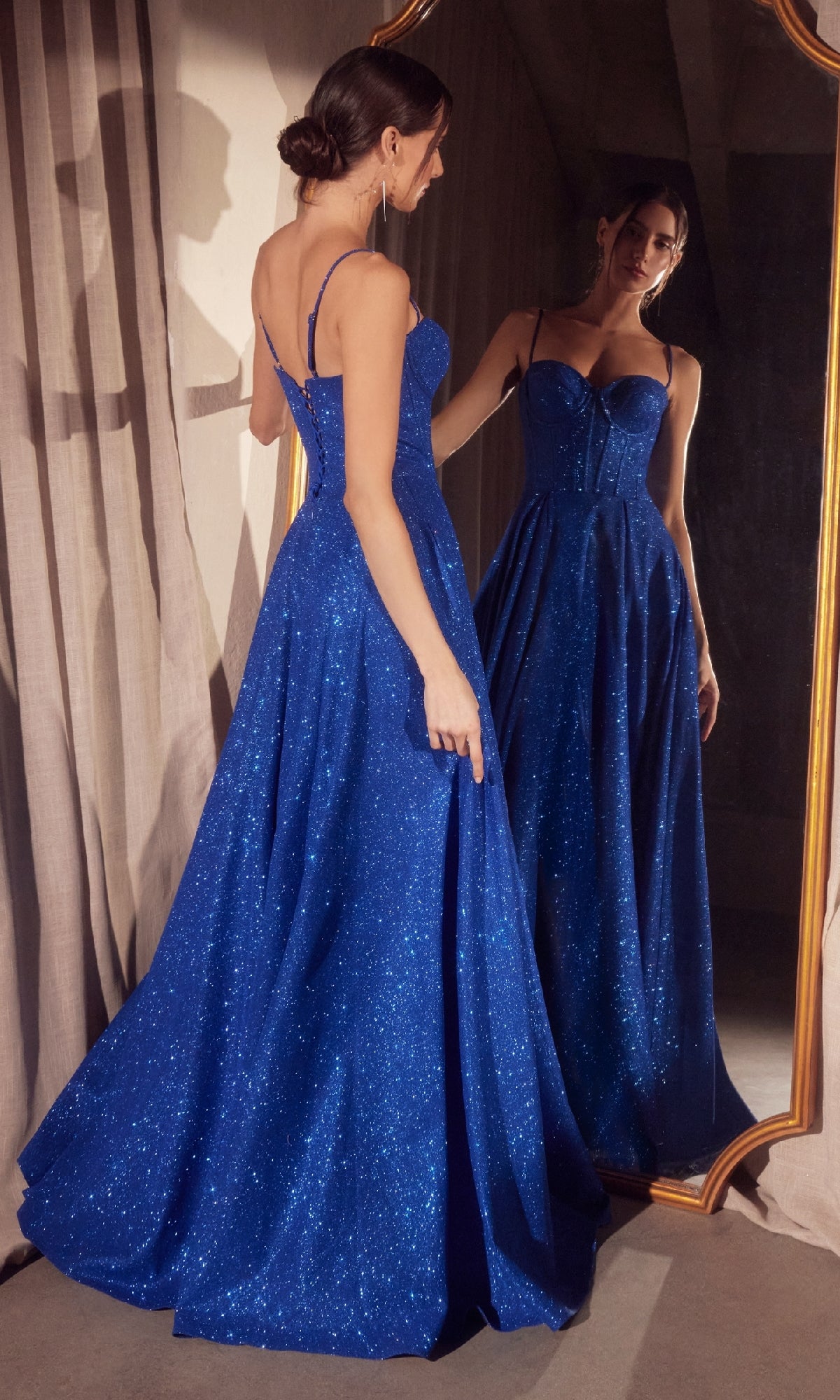 A woman in a sleeveless, sparkling blue Lace-Up Long Glitter Prom Ball Gown: Amoris T706 stands before a full-length mirror, admiring her reflection.